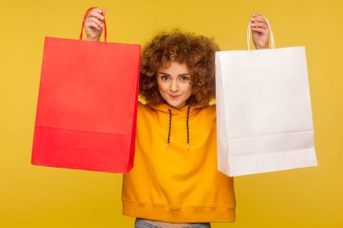 Portrait of happy shopper, curly-haired hipster girl in hoodie raising shopping bags with blank area, copy space on packages for advertising text. indoor studio shot, commercial of fashion store