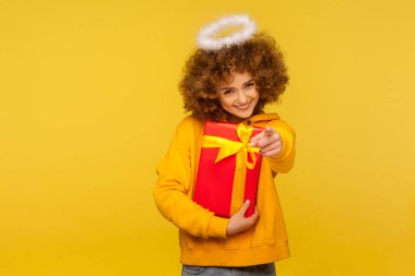 Hey you, happy holidays! Portrait of cheerful curly-haired angelic woman with saint nimbus pointing to camera and holding wrapped christmas gift box. indoor studio shot isolated on yellow background
