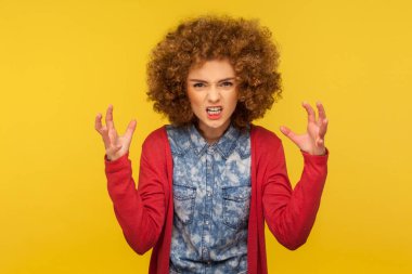Aggressive emotions. Portrait of crazy irritated woman with curly hair in casual outfit raising fist frustrated and furious, expressing anger, hate and rage. studio shot isolated on yellow background