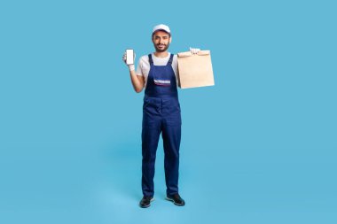 Full body portrait of expert courier in blue uniform holding groceries bag and mobile phone with mock up blank display for professional delivery advertise, online food purchase. studio shot isolated