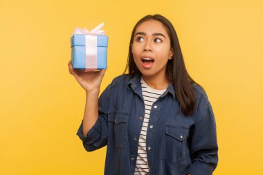 Portrait of funny amazed girl in denim shirt holding wrapped gift, looking up with open mouth and shocked expression, thinking what's inside box. indoor studio shot isolated on yellow background