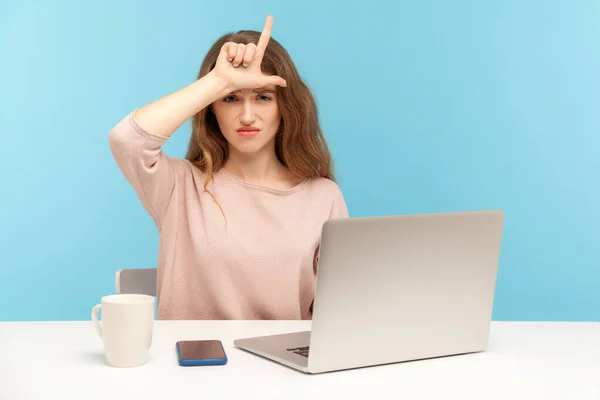 Lost job, unemployment. Depressed young woman employee sitting at workplace with laptop and showing loser gesture, desperate about unlucky dismissal. indoor studio shot isolated on blue background