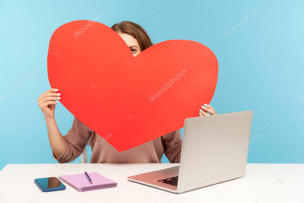Shy woman hiding face behind huge red paper heart and peeking curious, holding symbol of love, romantic feelings, using dating service on laptop, online chat and website. indoor studio shot isolated