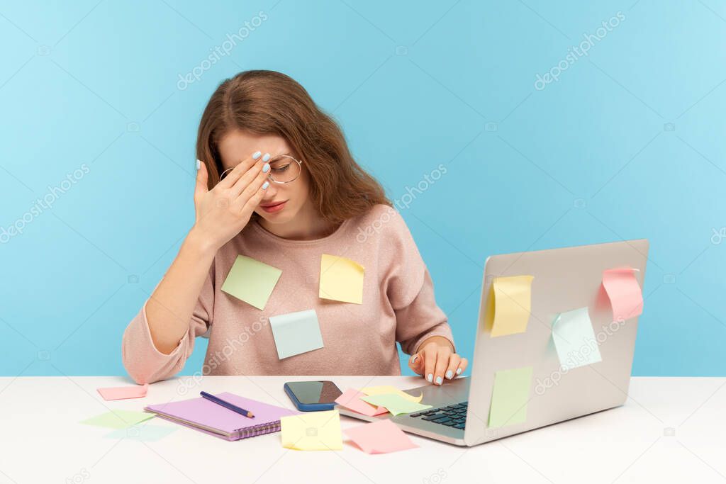 Exhausted fatigued overworked woman employee in glasses covered with sticky notes, touching head thinking desperate, suffering headache of work overload. indoor studio shot isolated on blue background
