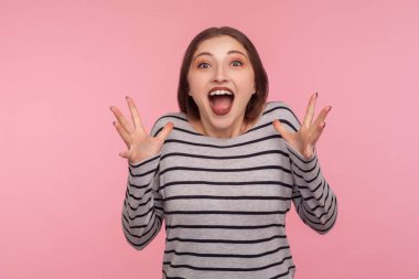 I can't believe this! Portrait of excited surprised woman in striped sweatshirt raising hands and shouting in amazement, shocked by sudden success. indoor studio shot isolated on pink background