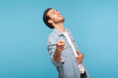 Ha ha, look at you! Portrait of man in worker denim shirt laughing out loud and pointing finger to camera, making fun of ridiculous appearance, amused by joke. studio shot isolated on blue background