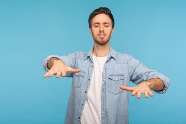 Portrait of blind unhappy man in denim shirt cautiously standing with closed eyes and touching air to find lost way, emulating walking in darkness. indoor studio shot isolated on blue background