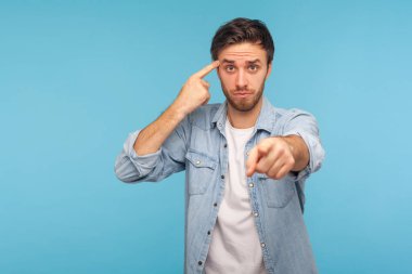 You are idiot! Portrait of man in denim shirt showing stupid gesture and pointing to camera, blaming for insane plan, crazy idea, dumb suggestion. indoor studio shot isolated on blue background