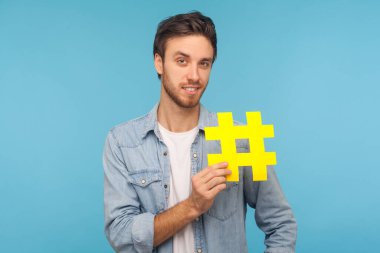 Portrait of cheerful man in denim shirt holding yellow hashtag symbol, standing isolated on blue background with copy space for tagged message, popular internet idea, viral content. studio shot