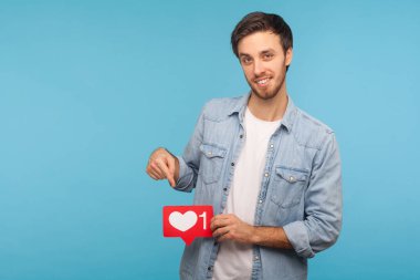 Click Like and subscribe! Portrait of handsome blogger man in denim shirt pointing at social media heart button, emoji counter, follower notification. indoor studio shot isolated on blue background