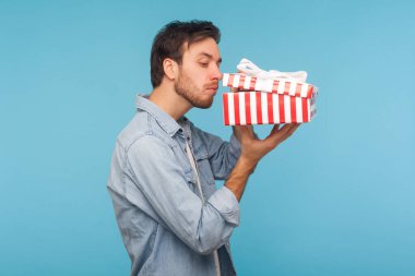 Portrait of curious man in denim shirt opening gift, peeking inside box with nosy look, unpacking present, in anticipation of interesting birthday surprise. studio shot isolated on blue background
