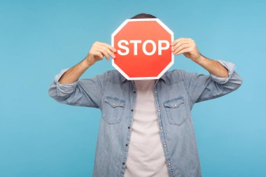 Portrait of anonymous man in worker denim shirt covering face with Stop road traffic sign as symbol of prohibition, restrictions and forbidden access. indoor studio shot isolated on blue background