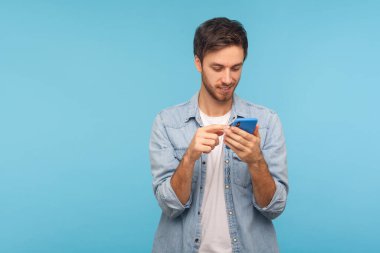 Portrait of happy smiling man in worker denim shirt texting in social media, reading positive message on cell phone, using mobile network services. indoor studio shot isolated on blue background