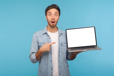 Portrait of amazed shocked man in worker denim shirt pointing at laptop screen with empty display, mock up free space for commercial image or text. indoor studio shot isolated on blue background