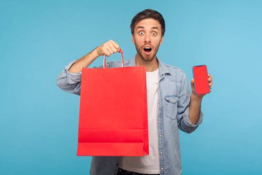 Amazed shocked man holding shopping bags and cell phone with mock up, blank display for advertisement of online store mobile app, goods purchase and delivery. studio shot isolated on blue background