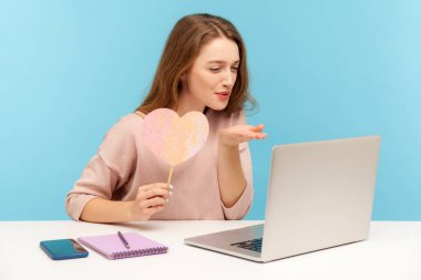 Pembe kalp tutan ve dizüstü bilgisayara hava öpücüğü gönderen, romantik duygular ifade eden, karantinada online randevusu olan bir kadın. Mavi arka planda kapalı stüdyo çekimleri