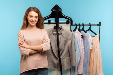 Kendine güvenen mutlu kadın, profesyonel moda tasarımcısı tasarımcı dükkanında raflarda asılı duran moda tasarımcısı, mağazada moda kıyafetleri satan. Mavi arka planda kapalı stüdyo çekimleri