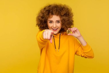 Hey you, buy digital money! Portrait of happy excited curly-haired young woman in urban style hoodie holding golden bitcoin and pointing to camera. indoor studio shot isolated on yellow background clipart