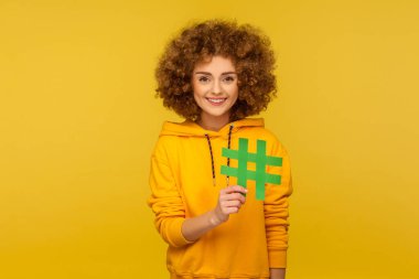 İnternet trendleri. Şehir tarzı kapüşonlu mutlu kıvırcık saçlı kadın portresi büyük hashtag işareti gösteriyor, blog tavsiyesi, viral gönderi etiketleme anlamına geliyor. Sarı arka planda izole edilmiş kapalı stüdyo çekimi