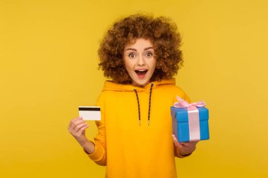 Shopping for electronic money. Portrait of surprised curly-haired young woman in urban style hoodie showing credit card and gift box, looking with amazement. studio shot isolated on yellow background clipart