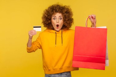 Alışveriş kredisi ve nakit para! Şok olmuş kıvırcık saçlı, kapüşonlu kızın portresi. Elinde kredi kartı ve reklam metni için içi boş paketler var. İç mekan stüdyo çekimi, moda mağazası reklamı.