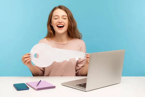 Excited emotions about house purchase. Amazing woman sitting at workplace with laptop, holding big paper key and laughing loudly, satisfied with new home, rental office. indoor studio shot isolated