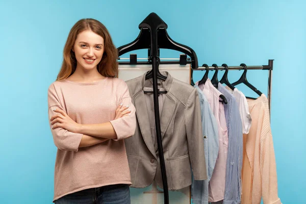 Confident happy woman, professional expert fashion stylist standing near clothes hang on shelf in designer store, selling trendy outfit in shop boutique. indoor studio shot isolated on blue background