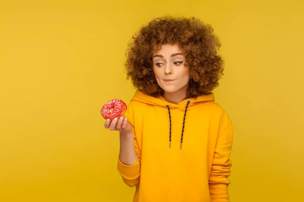 对垃圾食品的怀疑 穿着都市风格帽衫 体贴入微的卷发年轻女子的画像 带着困惑而不确定的表情望着甜甜圈 在黄色背景下隔离的室内拍摄 — 图库照片
