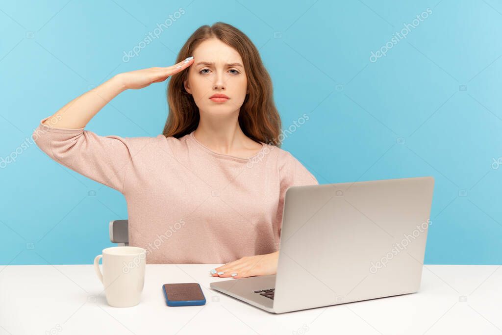 Yes sir! Obedient responsible young woman employee sitting at workplace with laptop and giving salute, listening to boss order, corporate discipline. indoor studio shot isolated on blue background