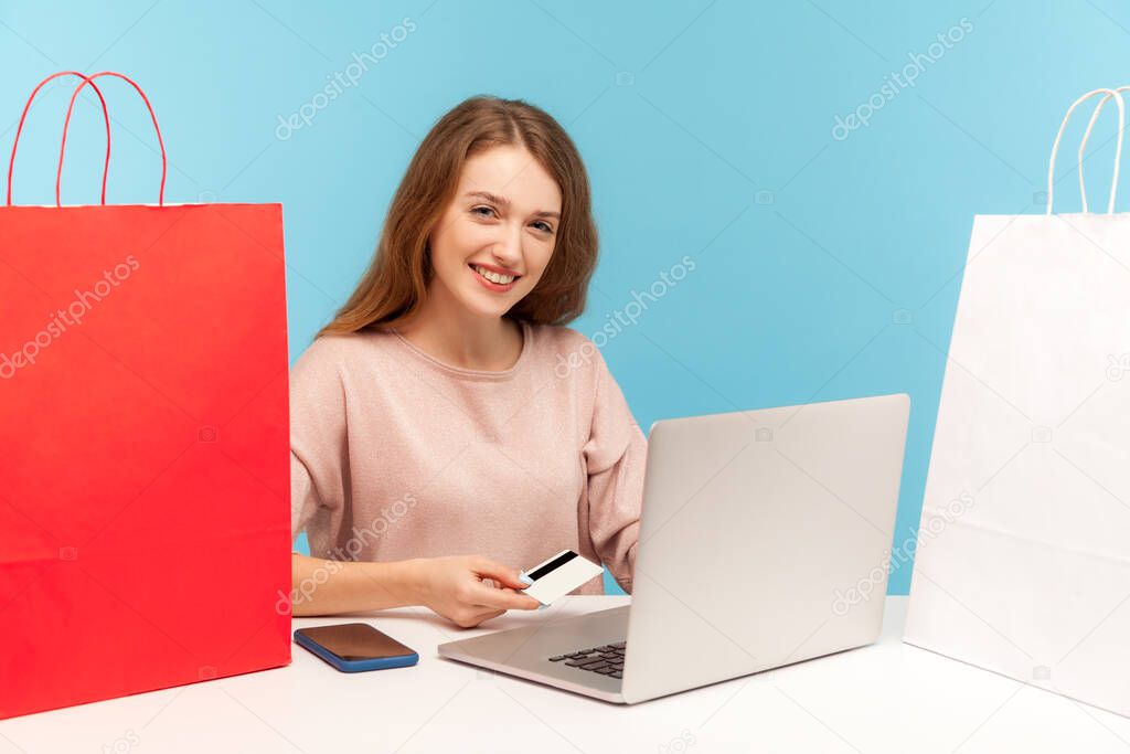 Happy young beautiful woman buyer surrounded by packages smiling, sitting at table shopping online on laptop, holding credit card to pay for purchase. indoor studio shot isolated on blue background