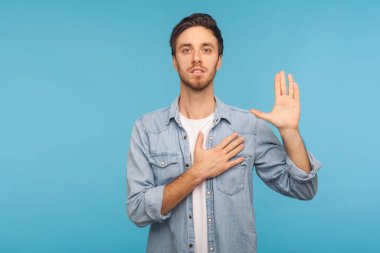 I swear! Portrait of honest responsible man in worker denim shirt giving evidence with raised hand as telling only truth, making loyalty promise oath. indoor studio shot isolated on blue background clipart