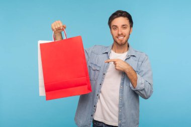 Kırmızı alışveriş torbalarını gösteren kot gömlekli yakışıklı esmer adamın portresi ve moda mağazasından alınmış paketleri gösteren gülümseme. Mavi arka planda kapalı stüdyo çekimleri
