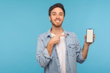 Çalışan kot gömlekli mutlu bir adamın portresi gülümseyen ve cep telefonu cihazını gösteren, cep telefonlu, reklam için boş bir ekran oluşturan. Mavi arkaplanda kapalı stüdyo çekimleri, kopyalama alanı