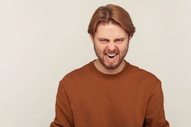 Tişörtlü sinirli ve sakallı bir adamın portresi kameraya karşı vahşice kükreyerek öfkeli, deli gibi bağıran ve nefret dolu. Gri arka planda kapalı stüdyo çekimi