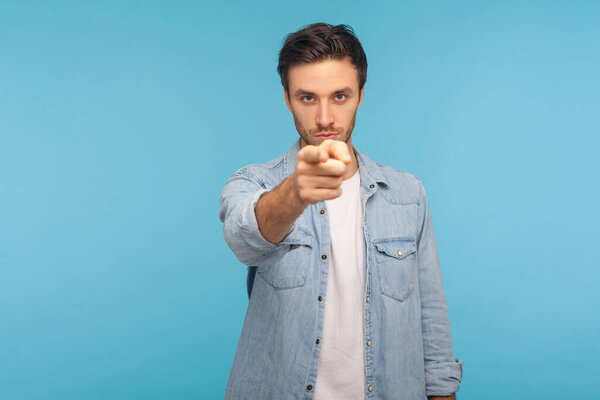 Hey you! Portrait of angry strict man in worker denim shirt noticing and pointing finger to camera, accusing with serious bossy face, making choice. indoor studio shot isolated on blue background