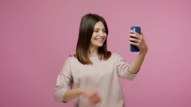 Güzel, genç, esmer bir kadın gülümsüyor, akıllı telefondan selfie çekerken eğleniyor, mobil uygulama kullanıyor, sosyal ağ paylaşımı için video çekiyor, blog yazıyor. Pembe arka planda kapalı stüdyo çekimleri