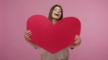 Sevgi ve enerji dolu neşeli esmer kız kocaman bir kalp taşıyor, kameraya gülüyor, dans ediyor, romantik duygular, ilgi ve cömertlik gösteriyor. Pembe arka planda kapalı stüdyo çekimleri