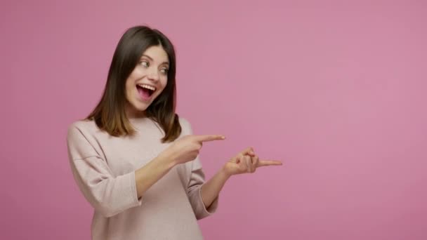 親切な笑顔で素晴らしいブルネットの女の子が親指を上げて空の場所 商業テキストの広告エリア 商品プロモーションのためのコピースペースを指しています ピンクの背景に隔離された屋内スタジオで — ストック動画