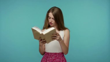 Yazlık elbise giyen, büyük kitaplar okuyan, ansiklopedi ya da dil sözlüğü okuyan kameraya gülümseyen ve baş parmak gösteren zeki kız öğrenci. Mavi arka planda kapalı stüdyo çekimleri