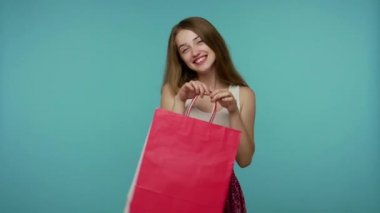 Yazlık elbiseli, elinde alışveriş poşetleri olan, gülümseyen, kaygısızca gülen, moda mağazasından neşe saçan, başparmağını kaldıran hoş bir kız. Jest gibi. Mavi arka planda kapalı stüdyo çekimleri