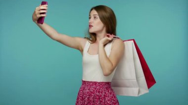 Elinde alışveriş poşetleri olan, cep telefonuna gülümseyen, selfie çeken ya da video görüşmesinde konuşan, moda mağazasından keyifli alışverişler yapan mutlu alışveriş kızı. İç mekan stüdyo çekimi, mavi arka plan