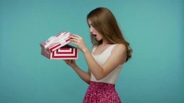 Yaz elbisesi içinde güzel bir kız hediyesini açıyor, kutuyu açıyor ve büyük bir sürpriz yapıyor, şaşırmış görünüyor, inanılmaz bir doğum günü hediyesi veriyor. Mavi arka planda kapalı stüdyo çekimleri