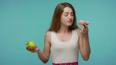 Sıska, güzel bir kız donut kokluyor ve elma yiyor, lezzetli meyveli ve şekerli çörekler, vitamin beslenme konsepti ve sağlıklı beslenme arasında seçim yapıyor. Stüdyo çekimi mavi arkaplanda izole edildi