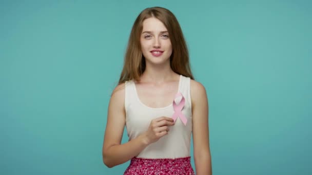 穿着夏装的可爱善良的女孩 带着粉红缎带 对着相机微笑 警告着乳腺癌的风险 女性的保健观念 每年的诊断 室内拍摄 蓝色背景隔离 — 图库视频影像