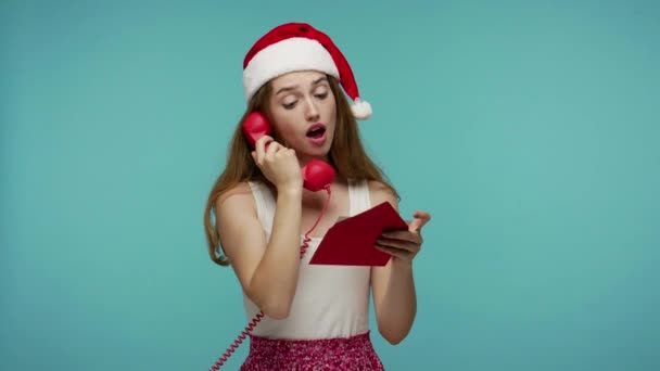 圣诞节快乐可爱的圣诞小精灵女孩 戴着圣诞礼帽 阅读信袋和礼物清单 在手机上交谈 澄清订单 圣诞节的信息 室内拍摄 蓝色背景 — 图库视频影像