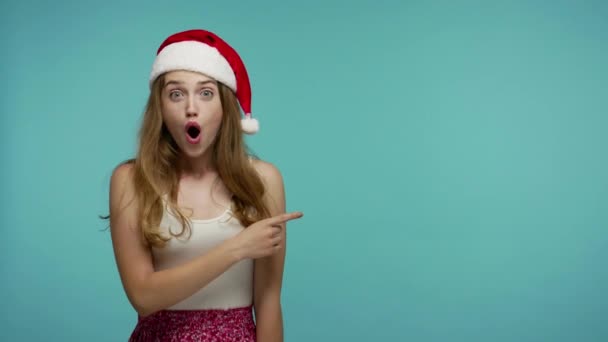 头戴滑稽圣诞礼帽的女孩惊奇地指着空白的广告区 惊讶地看着空旷的地方 寻找商业形象或圣诞礼物清单上的文字 新年祝福 被隔离的工作室 — 图库视频影像