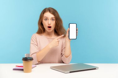 Şaşırmış bir kadın, ofis çalışanı, iş yerinde oturan ve cep telefonunu gösteren bir görüntü, başvuru için kopya alanı, web sitesi reklamı. Mavi arka planda kapalı stüdyo çekimleri