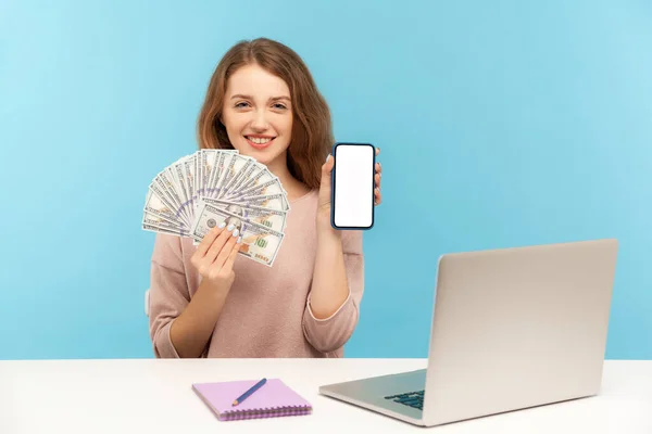 Freelance job, internet earnings online. Young positive woman sitting at workplace and holding dollar banknotes, showing smartphone with mockup display. indoor studio shot isolated on blue background