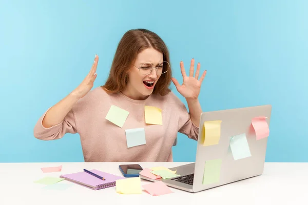 Depressed, stressed out, overworked woman employee in glasses covered with sticky notes, angrily raising hands, screaming crazy in desperate at laptop. indoor studio shot isolated on blue background
