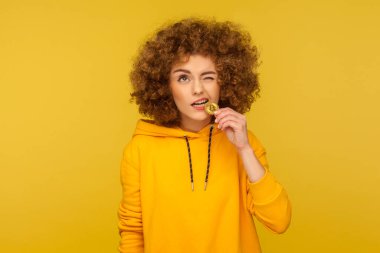 Kripto para birimini deneyelim! Kıvırcık saçlı kapüşonlu genç bir kadının portresi altın bitcoin ısırıyor, dişinde btc madeni para tadıyor, zincir kavramı. Stüdyo çekimi sarı arkaplanda izole edildi